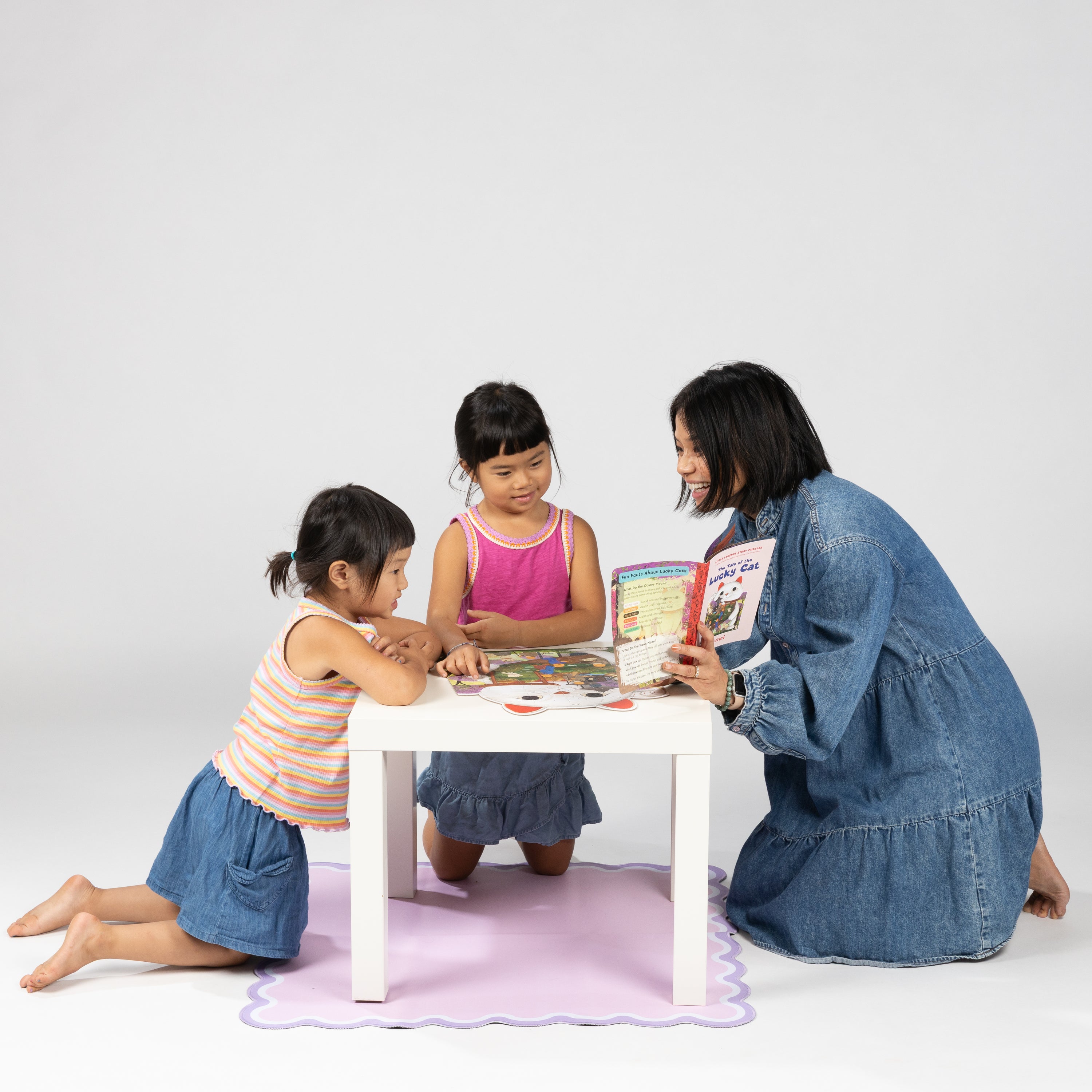Three children sitting around a small table with a book, on a white background, doing a jigsaw puzzle and reading a story